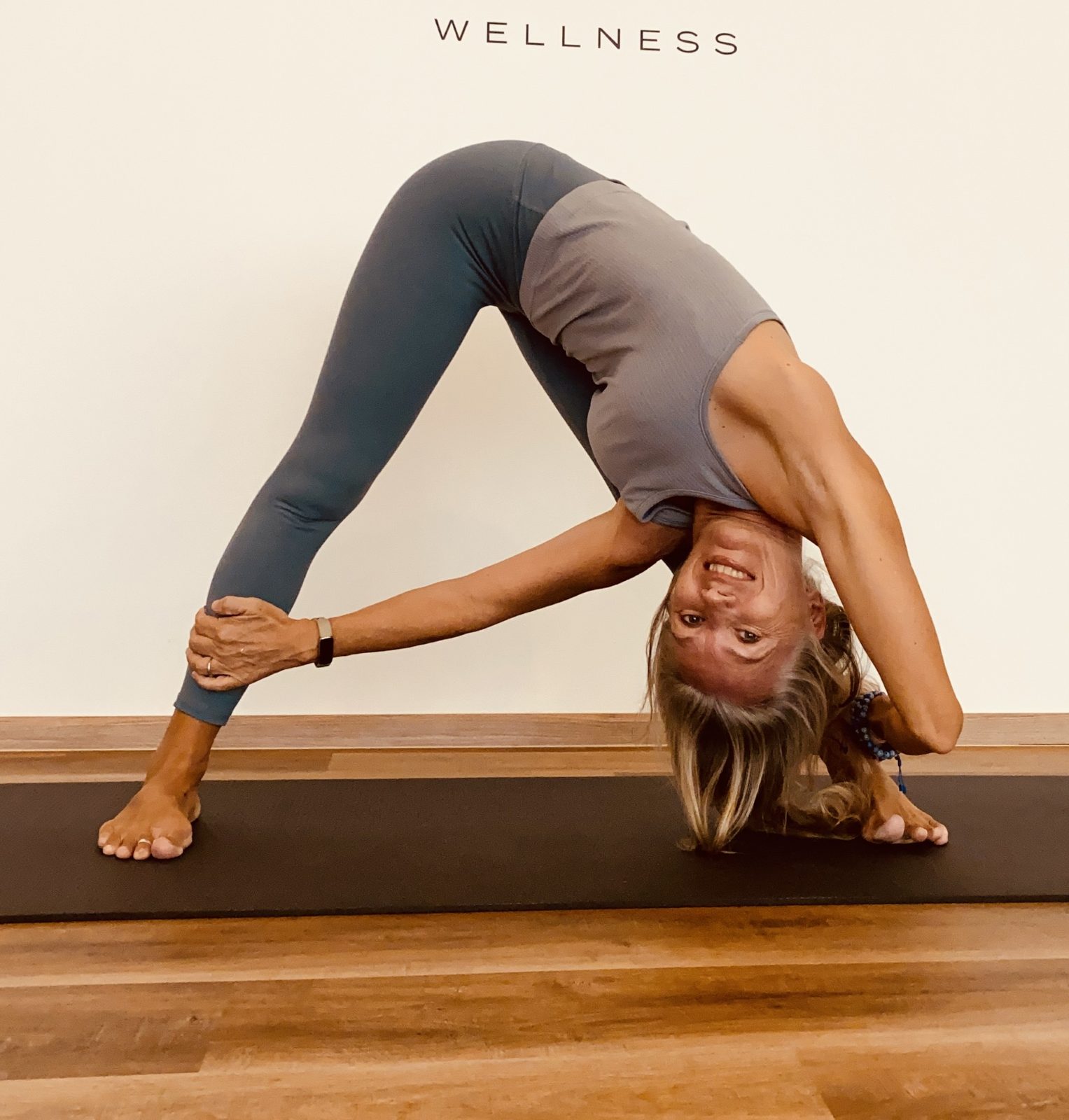 Laurence smiling in an upside-down downward dog / wheel stretch on a mat, with a "Wellness" sign on the wall behind her