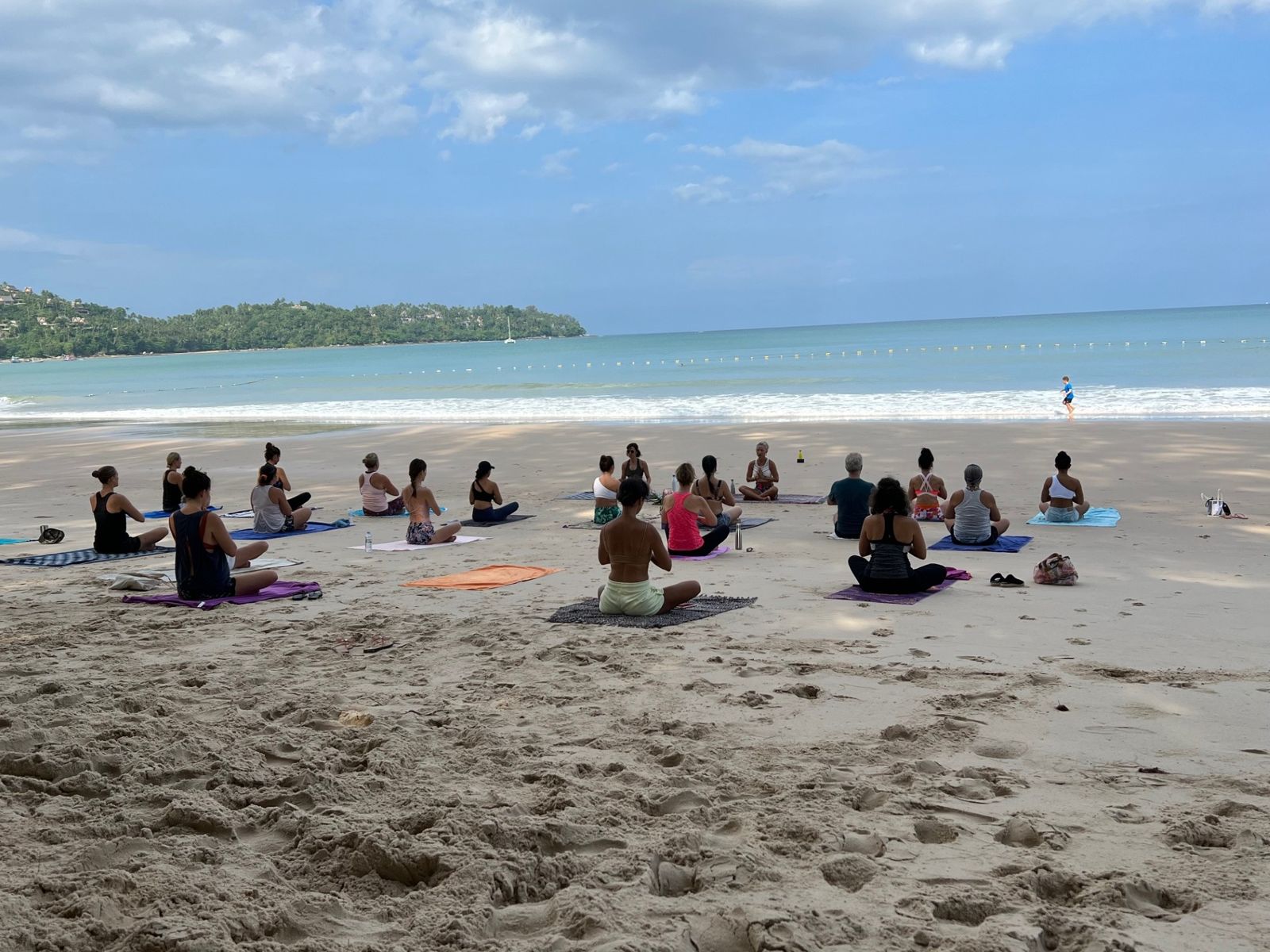 A group seated in meditation on the sand facing Laurence, with the sea and a green headland behind