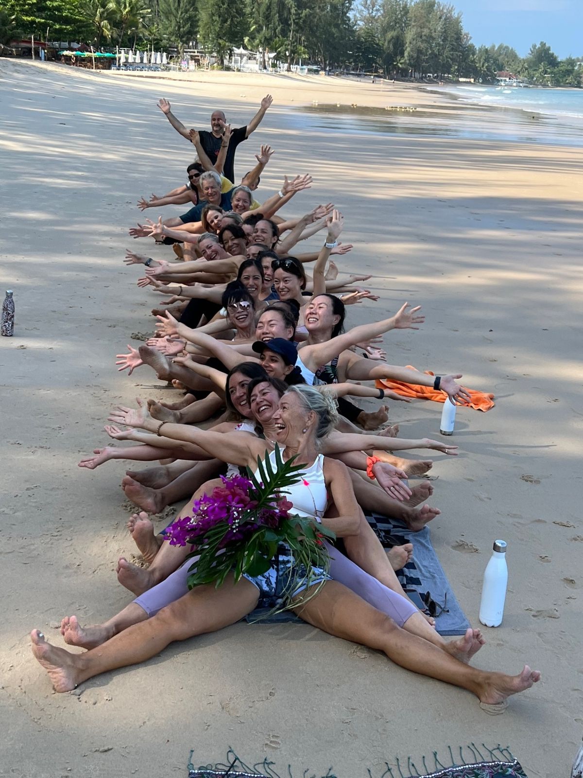 A laughing group sitting in a long line on the beach with arms stretched out, Laurence at the front